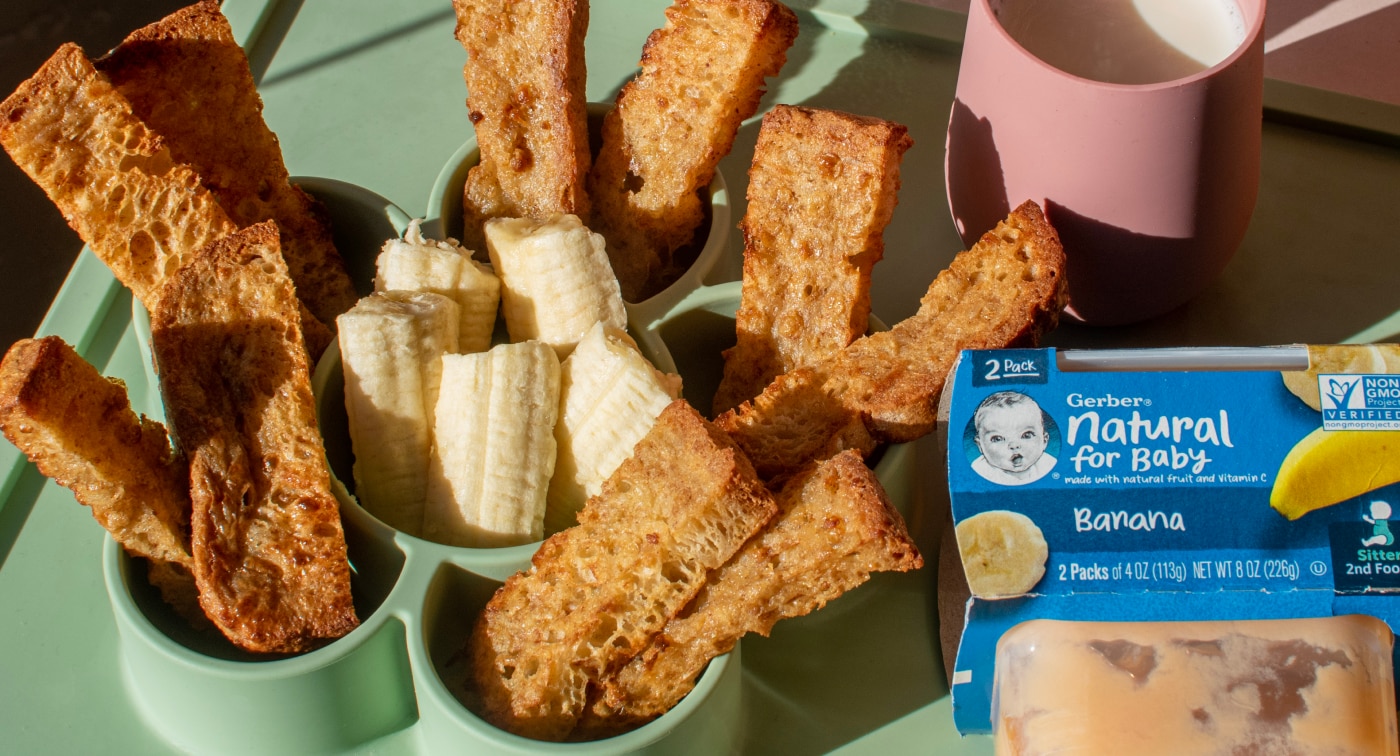 Batch of banana french toast sticks made with Gerber banana puree on a plate with cut up banana and a glass of milk.