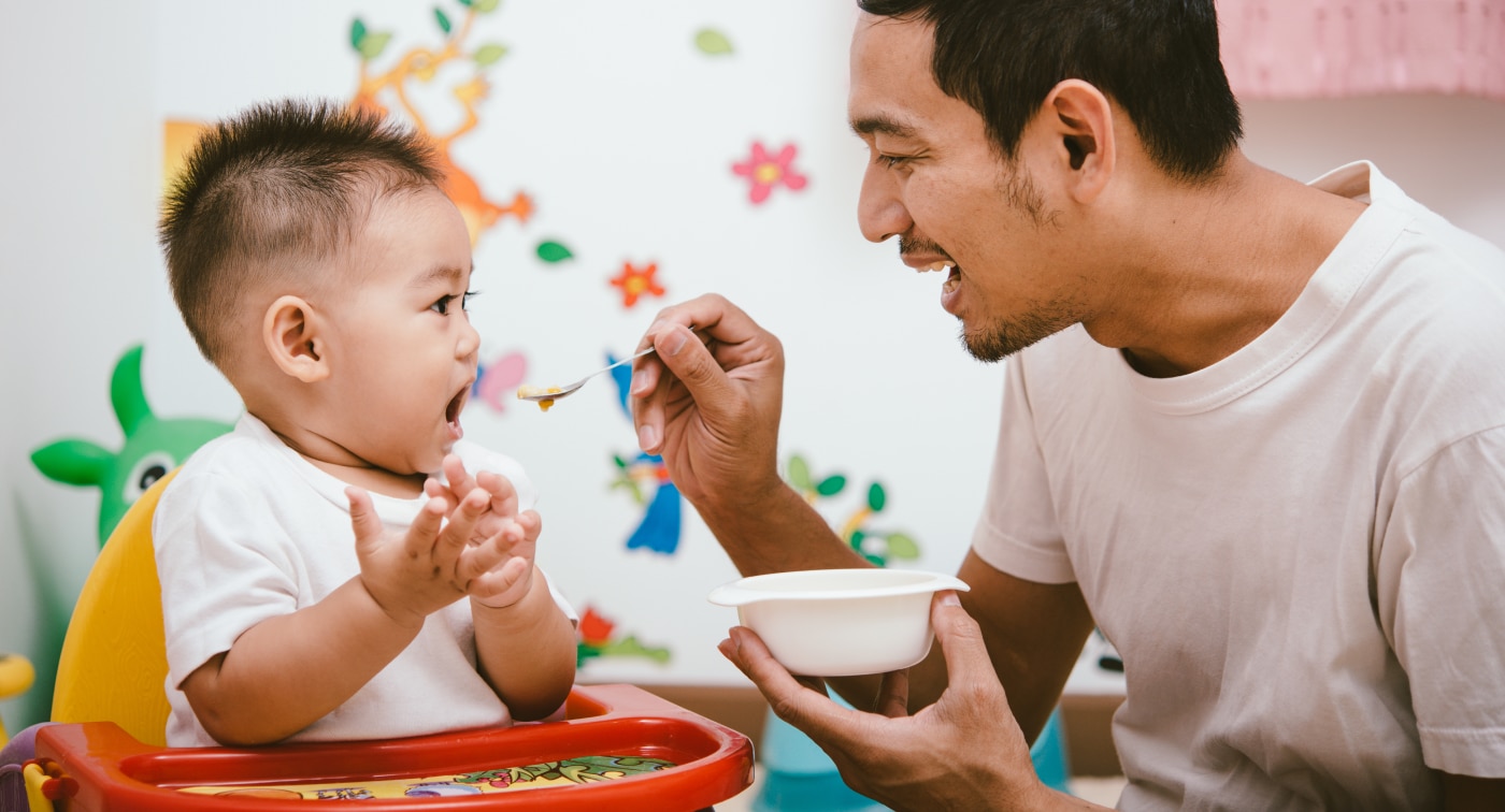 Dad holding a bowl of food and spoon-feeding his toddler sitting in a highchair.