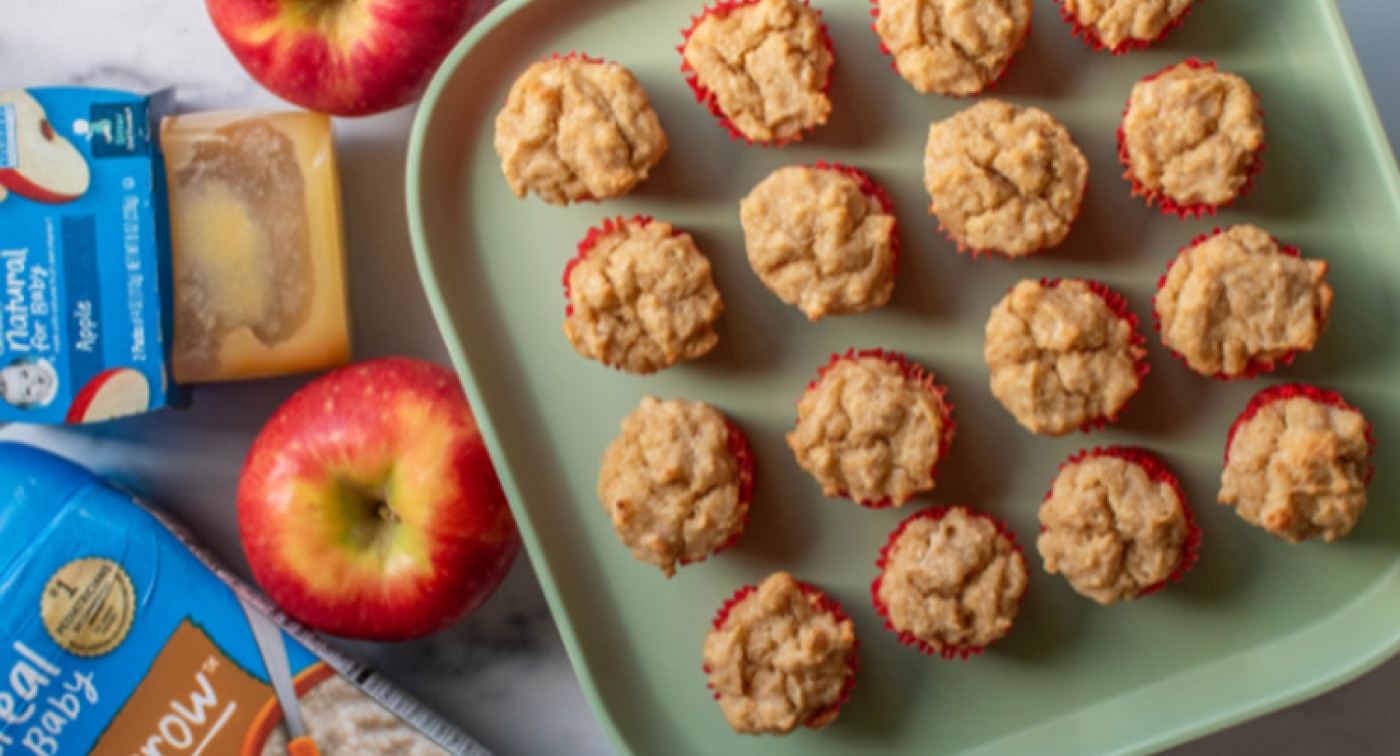 One dozen baked apple cinnamon mini muffins made with Gerber oatmeal cereal and Gerber apple puree and arranged on a plate surrounded by fresh apples.